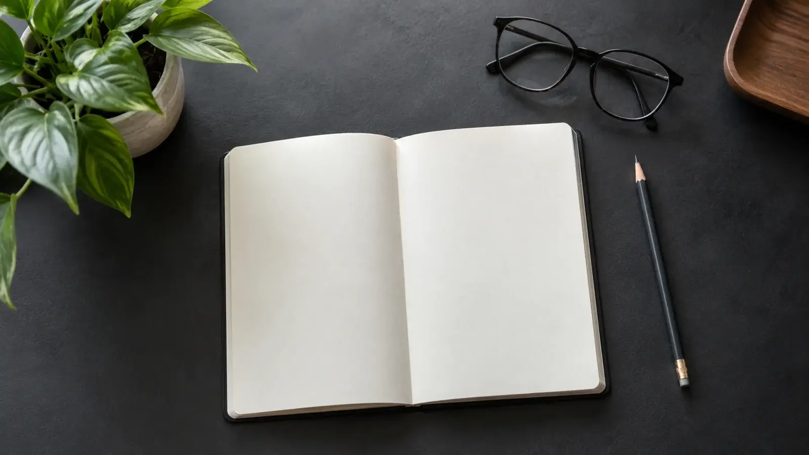 A notebook, glasses, pencil, and plant sit on a charcoal desk.