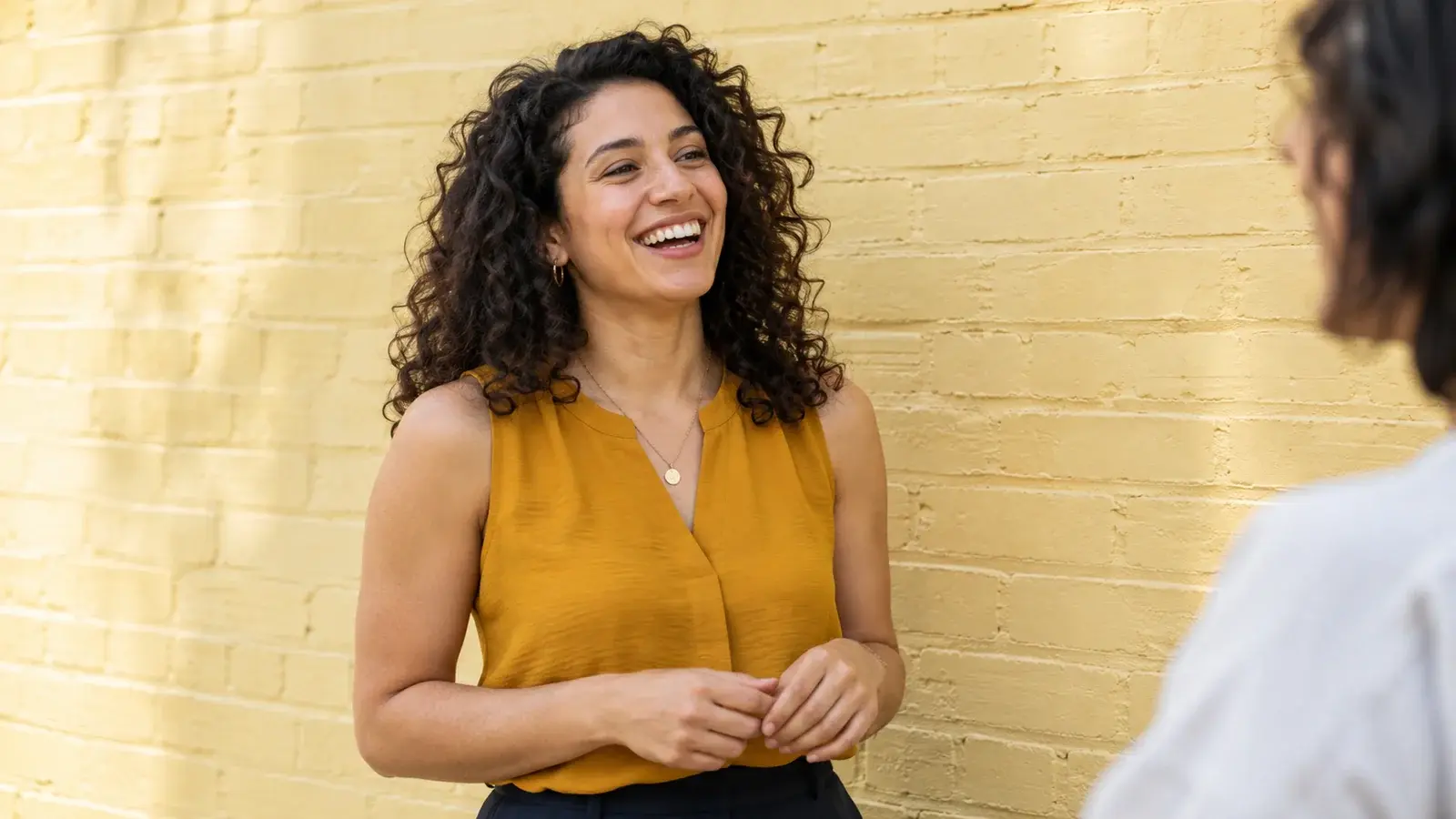 A woman with curly hair laughs during a conversation outdoors.