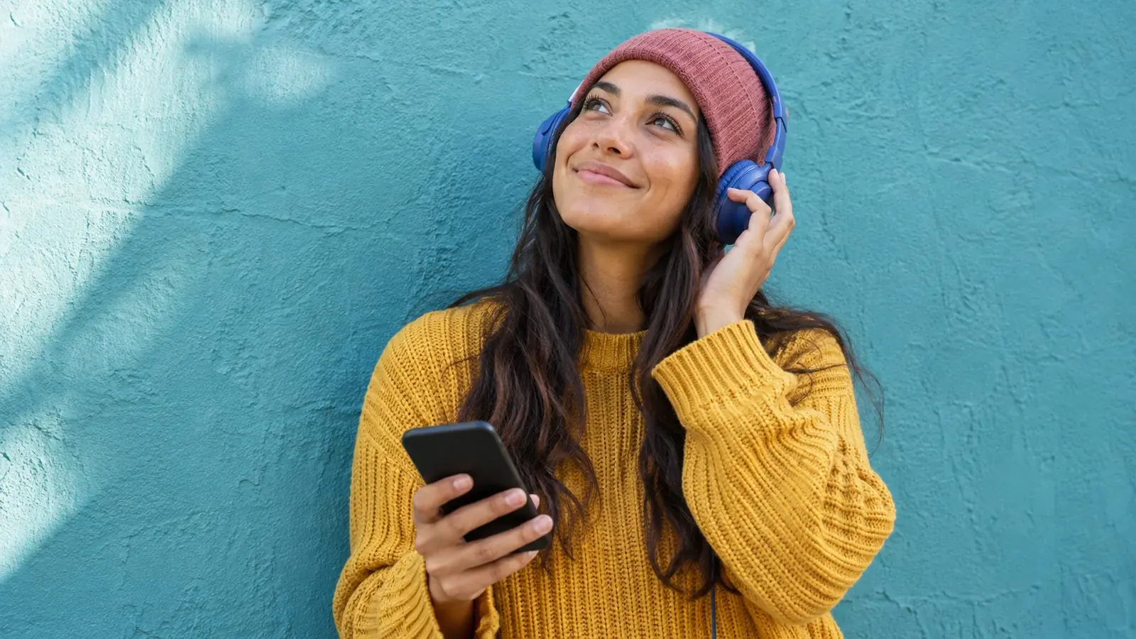 A woman in a yellow sweater listens through headphones while holding her phone.