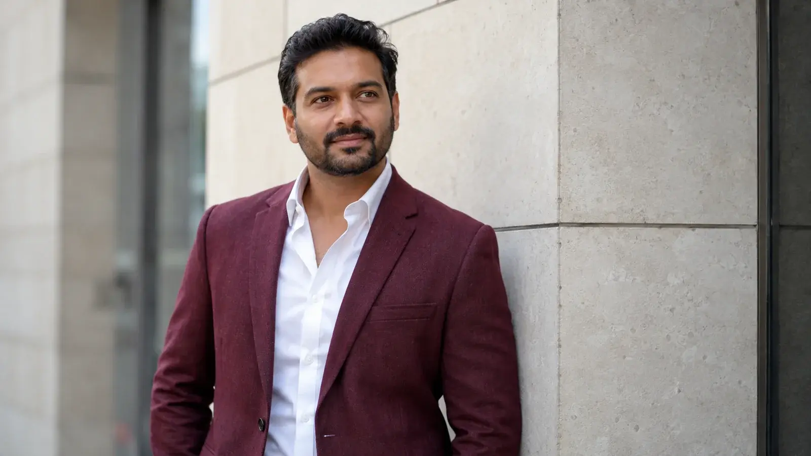 A confident sales professional in a burgundy blazer stands near an office wall.